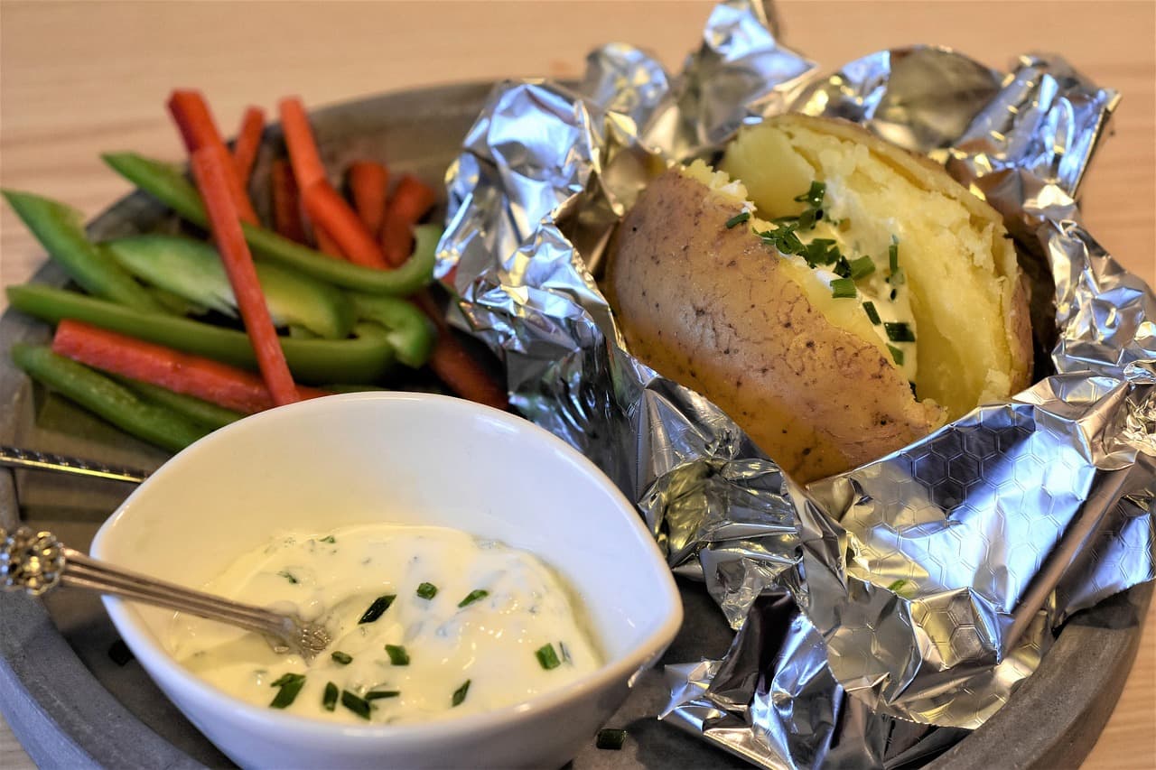 A jacket potato split open in foil, with a pot of soured cream and chives